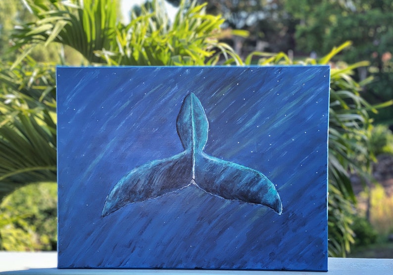Puede incluir: Una pintura sobre lienzo representa la cola de una ballena en tonos azules y turquesas sobre un fondo azul oscuro con puntos blancos en forma de estrellas. La obra de arte est&aacute; situada sobre un fondo de follaje verde.