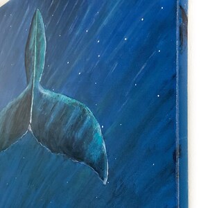 Puede incluir: Una pintura sobre lienzo representa la cola de una ballena en tonos azules sobre un fondo azul oscuro con estrellas blancas. La obra de arte se ve desde un &aacute;ngulo lateral, mostrando el lienzo texturizado y el borde de la pintura.