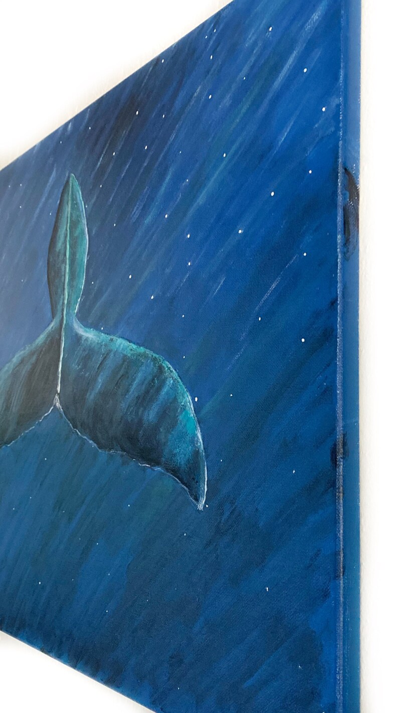 Puede incluir: Una pintura sobre lienzo representa la cola de una ballena en tonos azules sobre un fondo azul oscuro con estrellas blancas. La obra de arte se ve desde un &aacute;ngulo lateral, mostrando el lienzo texturizado y el borde de la pintura.