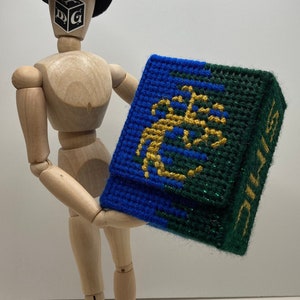 May include: A wooden mannequin wearing a black hat with a dice graphic on the front, holding a blue and green fabric dice bag with a gold embroidered design. The bag has the word "DICE" on the side.