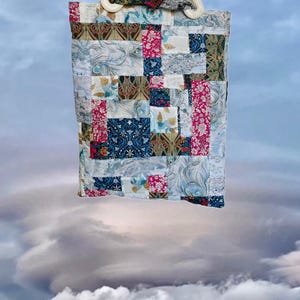 Può includere: Una borsa tote patchwork con manico color crema. La borsa presenta vari motivi di tessuto nei toni del blu, rosa e verde. Il manico è in plastica curva bianco sporco. La borsa è sospesa contro un cielo nuvoloso.