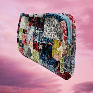 May include: A patchwork fabric zippered pouch with a variety of floral and geometric patterns in shades of blue, green, pink, and white. The pouch is floating against a pink and purple cloudy sky.