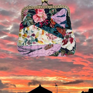 Op de afbeelding: Een bloemen patchwork clutch met een messing sluiting. De tas heeft een verscheidenheid aan kleurrijke bloempatronen in roze, wit en blauw. Het frame heeft een donker bronzen kleur. De achtergrond is een zonsondergang.