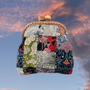 Op de afbeelding: Een patchwork clutch met een messing frame en houten sluiting. De stof is een mix van bloemen- en patroonontwerpen in verschillende kleuren, waaronder rood, groen en blauw. De tas staat tegen een luchtachtergrond.