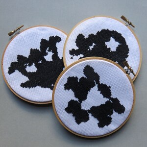 May include: Three round embroidery hoops with white fabric and black ink designs. The designs are abstract and resemble ink blots.