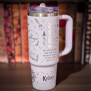 May include: A light pink Stanley tumbler with a handle and clear lid, featuring black line art of a sword, quill, book, and stars. The text reads Always, the name Kelsey, and You will also find that help will always be given to those who ask for it.