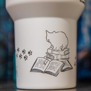 A white ceramic mug with a black and white illustration of a cat sitting on a stack of books.