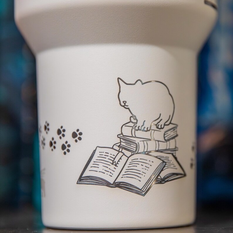 A white ceramic mug with a black and white illustration of a cat sitting on a stack of books.