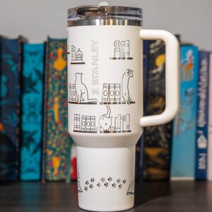 A white Stanley tumbler mug with a handle, featuring various illustrations of books and cats on its surface.