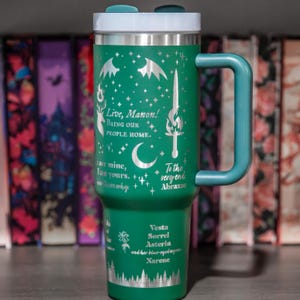 A green travel mug with a handle, featuring a design with stars, a sword, and text that appears to be a quote or message.