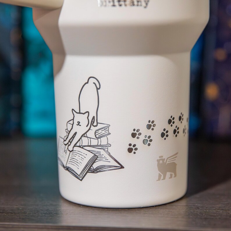 A white mug with a black and white illustration of a cat sitting on a stack of books, along with paw prints and a winged cat symbol.