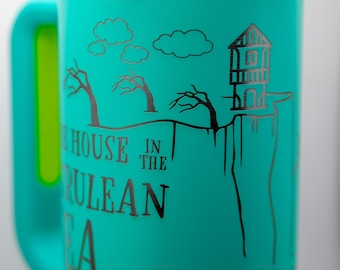 May include: A teal stainless steel tumbler with a handle, featuring a black laser-engraved illustration of a house on a cliff, bare trees, and clouds. The text reads "THE HOUSE IN THE CERULEAN SEA" in a serif font. The rim is polished silver metal.