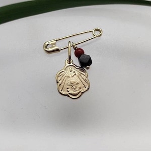 May include: A gold safety pin charm with a religious medal, a black bead, and a red bead.