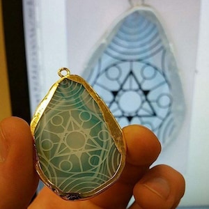 Könnte beinhalten: Ein goldfarbener Anhänger mit einem blauen Achat-Stein, der mit einem weißen geometrischen Muster verziert ist. Der Anhänger hat die Form einer Träne.