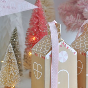Puede incluir: Dos cajas de regalo de color marrón con forma de casa de jengibre con cinta de lunares rosa y blanca. Las cajas tienen detalles blancos y están decoradas con diseños de bastones de caramelo y bombillas. Las cajas están sobre una superficie blanca con una cadena de luces blancas.