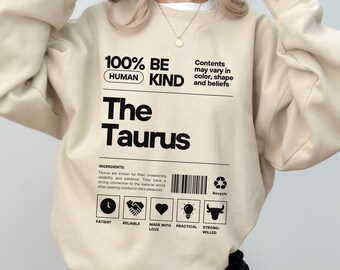 May include: A beige sweatshirt with black text that reads "100% Be Human Kind The Taurus". The text is surrounded by a black rectangle with the words "Contents may vary in color, shape and beliefs" above it. Below the rectangle is a black and white graphic with the words "Ingredients: Taurus are known for their unwavering reliability and patience. They have a strong connection to the material world often seeking comfort in life's pleasures." Below the graphic are six black and white icons with the words "Patient, Reliable, Made with Love, Practical, Strong-Willed" below them.