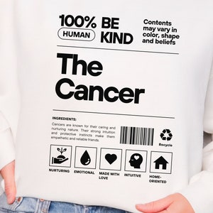 May include: White sweatshirt with a black graphic design that says "100% Human Be Kind The Cancer". The graphic is styled like a product label with a barcode, recycle symbol, and icons representing nurturing, emotional, made with love, intuitive, and home-oriented.