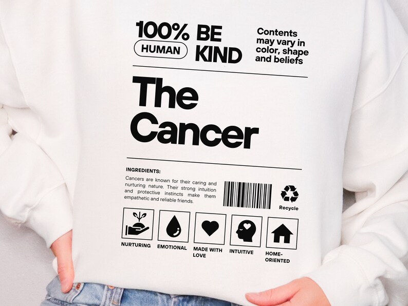 May include: White sweatshirt with a black graphic design that says "100% Human Be Kind The Cancer". The graphic is styled like a product label with a barcode, recycle symbol, and icons representing nurturing, emotional, made with love, intuitive, and home-oriented.