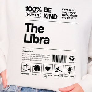 May include: White sweatshirt with black text that reads "100% BE HUMAN KIND The Libra". The text is in a product label format with a barcode and a recycle symbol. The label also includes the text "Ingredients: Libras are natural peacemakers. With their charming and sociable personality, they excel in bringing people together and fostering a sense of unity." Below the text are five icons: a scale, three people holding hands, a heart, a gavel, and a lightbulb. The icons are labeled "Balanced", "Social", "Made with Love", "Diplomatic", and "Intelligent".