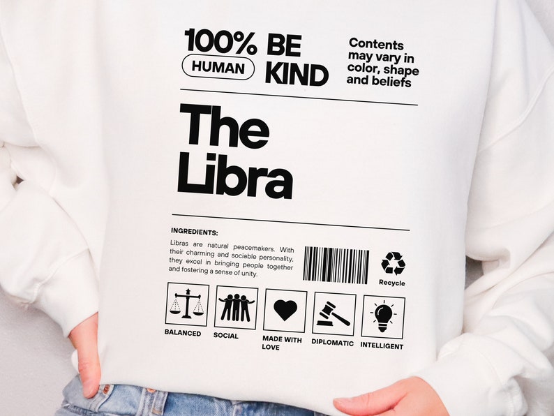 May include: White sweatshirt with black text that reads "100% BE HUMAN KIND The Libra". The text is in a product label format with a barcode and a recycle symbol. The label also includes the text "Ingredients: Libras are natural peacemakers. With their charming and sociable personality, they excel in bringing people together and fostering a sense of unity." Below the text are five icons: a scale, three people holding hands, a heart, a gavel, and a lightbulb. The icons are labeled "Balanced", "Social", "Made with Love", "Diplomatic", and "Intelligent".