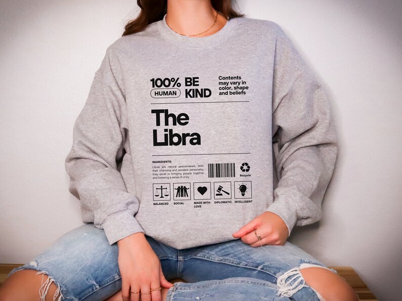 May include: Gray sweatshirt with black text that reads "100% Be Human Kind The Libra". The text is in a box with a barcode and symbols representing balanced, social, made with love, diplomatic, and intelligent.