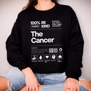 May include: Black sweatshirt with white text that reads "100% Be Human Kind The Cancer". The text is in a box with a barcode and icons that represent nurturing, emotional, made with love, intuitive, and home oriented.