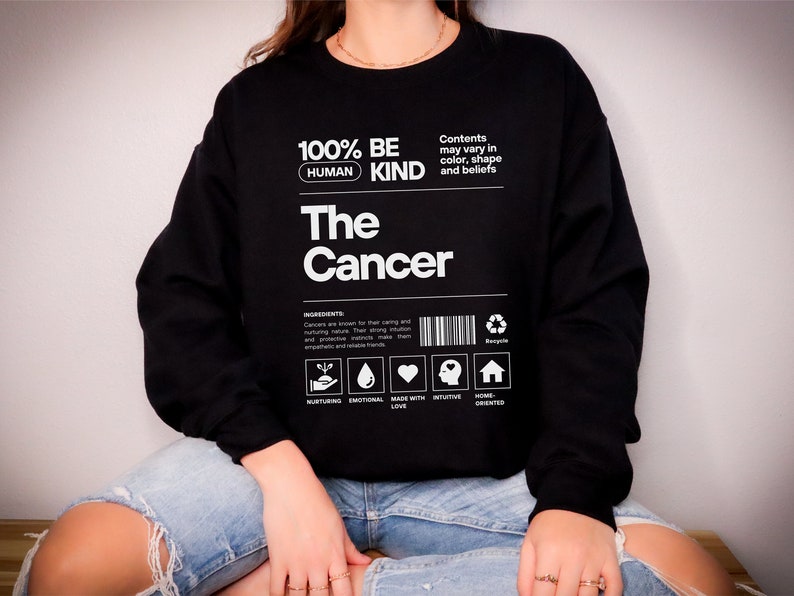 May include: Black sweatshirt with white text that reads "100% Be Human Kind The Cancer". The text is in a box with a barcode and icons that represent nurturing, emotional, made with love, intuitive, and home oriented.