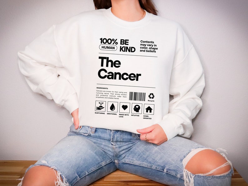 May include: White sweatshirt with black text that reads "100% Human Be Kind The Cancer". The text is styled to look like a product label with a barcode and icons.