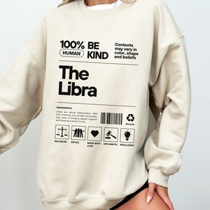 May include: A beige sweatshirt with black text that reads "100% BE HUMAN KIND The Libra". The text is in a label format with a barcode and a list of characteristics: Balanced, Social, Made with Love, Diplomatic, Intelligent.