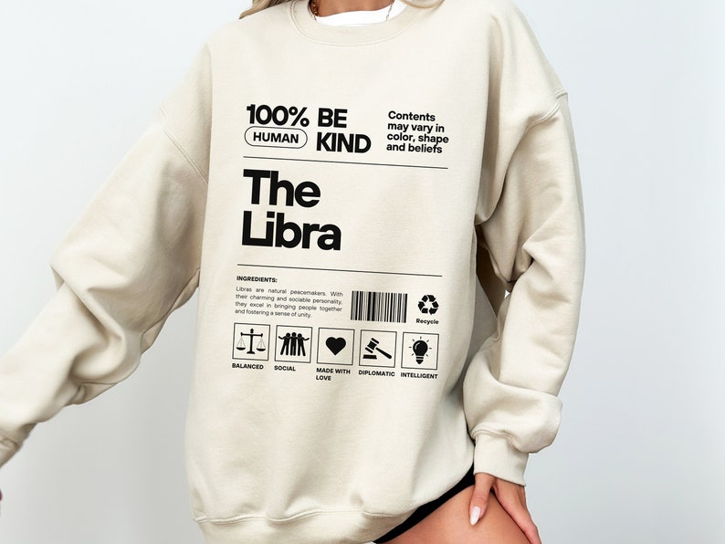 May include: A beige sweatshirt with black text that reads "100% BE HUMAN KIND The Libra". The text is in a label format with a barcode and a list of characteristics: Balanced, Social, Made with Love, Diplomatic, Intelligent.