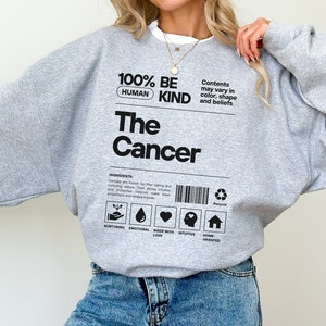 May include: Gray sweatshirt with the text "100% Human Be Kind The Cancer" and a label with icons and text that reads "Contents may vary in color, shape and beliefs".