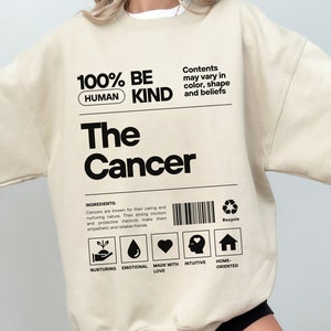 May include: A beige sweatshirt with black text that reads "100% Be Human Kind" and "The Cancer". The text is in a box with a barcode and a recycle symbol. The text below the box reads "Ingredients: Cancers are known for their caring and nurturing nature. Their strong intuition and protective instincts make them empathetic and reliable friends." There are five icons below the text: a hand holding a plant, a water drop, a heart, a head with a heart, and a house.