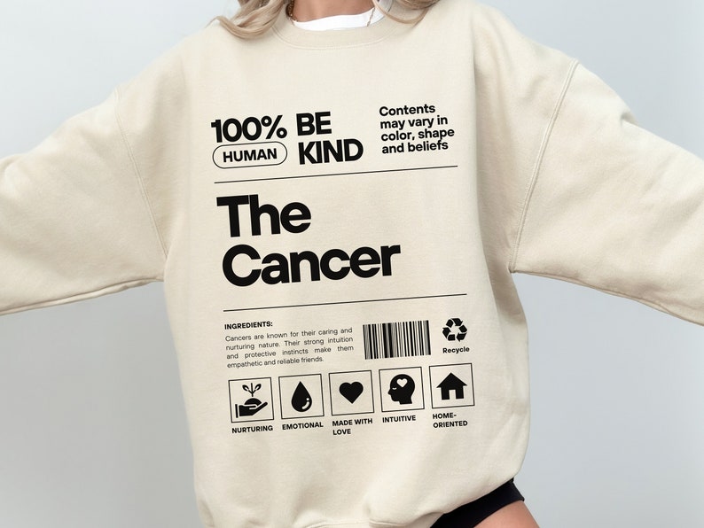 May include: A beige sweatshirt with black text that reads "100% Be Human Kind" and "The Cancer". The text is in a box with a barcode and a recycle symbol. The text below the box reads "Ingredients: Cancers are known for their caring and nurturing nature. Their strong intuition and protective instincts make them empathetic and reliable friends." There are five icons below the text: a hand holding a plant, a water drop, a heart, a head with a heart, and a house.