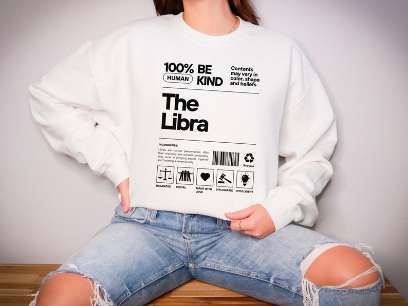May include: White sweatshirt with black text that reads "100% Human Be Kind The Libra". The text is styled to look like a product label with a barcode and icons representing balanced, social, made with love, diplomatic, and intelligent.