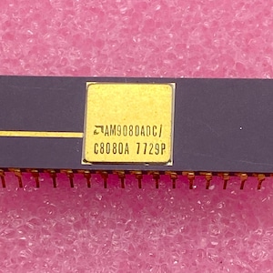 May include: A rectangular, dark gray integrated circuit with gold pins. A gold square in the center features the text "AM9080ADC/C8080A 7729P". The component is set against a pink, textured background.