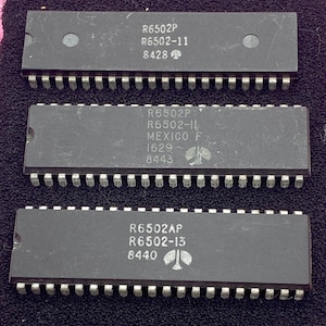 May include: Three black integrated circuit chips with silver pins. Each chip has text including "R6502" and other numbers. The chips are on a dark pink surface.