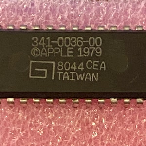 May include: A black rectangular integrated circuit chip with silver pins along the top and bottom edges. The surface features white text reading 341-0036-00, ©APPLE 1979, a square logo, 8044 CEA, and TAIWAN. The component has a circular indentation on the left.
