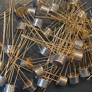 May include: A collection of numerous metal electronic transistors featuring cylindrical silver-coloured bodies and multiple thin, gold-coloured wire leads extending from the base. Each component displays small printed text on the top surface.