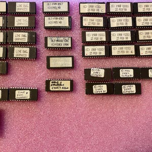 May include: A collection of black rectangular integrated circuit chips with white labels. The labels feature text including LINE DWG. GRAPHICS, ULT-FRM-BSC7 (C)1985 HD, (C) 1980 VIDEX 23 FEB 88, APPLE II ADAPTALK (C)726-HIGH, and 4-S KEYBOARD 5:30.