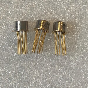 May include: Three electronic components with cylindrical metal tops and multiple thin gold-colored pins extending downwards. Each component features printed text including 723BE02 and a logo on the side of the metallic casing.