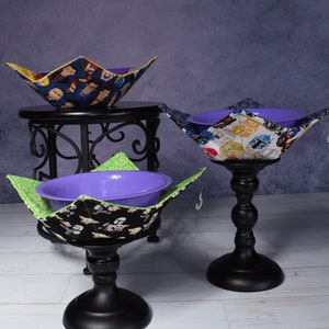 May include: Three fabric bowl cozies featuring various character prints, each holding a purple plastic bowl. The cozies display patterns of figures in blue, white, and black fabrics, with one interior lining in bright green and another in yellow.