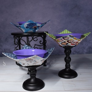 May include: Three fabric-covered bowls with purple, blue, and green patterns. The bowls are on black stands and are filled with purple bowls.
