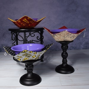 May include: Three fabric bowl cozies with different patterns and colors. The cozies are on black stands. The cozies are designed to hold bowls and keep them warm or cold.