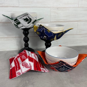 May include: A collection of bowl cozies in various patterns and colors. One cozy features the Michigan State Spartans logo, another the Detroit Red Wings logo, and others with navy and orange designs. The cozies are designed to hold bowls.
