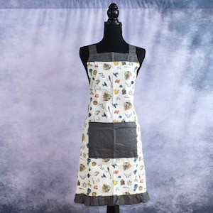 May include: A white apron with a gray pocket and a colorful print of various gardening tools and plants. The apron has a ruffled hem.