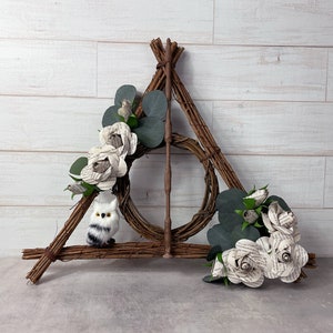 May include: A handcrafted, brown twig Deathly Hallows symbol with a wand in the center. The symbol is decorated with paper roses, green leaves, and a small, fluffy owl. The background is a white wooden wall.