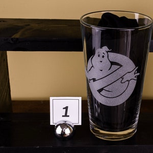 May include: A clear glass pint glass with a white etched design of the Ghostbusters logo and a ghost. The glass is sitting on a black shelf with a number 1 tag.