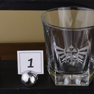 May include: A clear, etched glass tumbler featuring a stylized emblem. The glass has a square base and a slightly tapered design. A small white card with the number "1" is displayed in front of the glass, held by a silver metal holder.