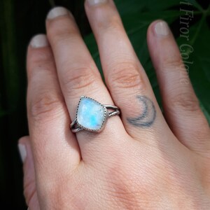 May include: A silver ring with a white moonstone set in a square shape. The ring is on a person's finger.