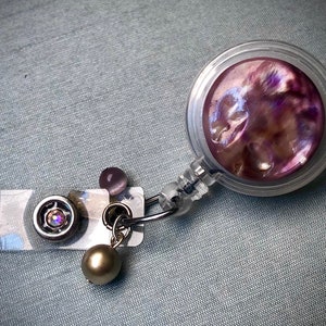 May include: A clear plastic retractable badge holder with a purple iridescent shell design. The holder has a silver swivel clip and a small pearl-like charm.